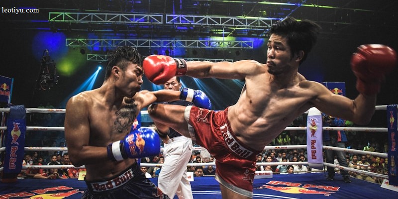 Two athletes are engaged in a dynamic kickboxing match inside a ring, surrounded by a cheering audience. One fighter, wearing red gloves and shorts, executes a high kick towards his opponent, who attempts to block the attack with his arm. The background features colorful lighting and promotional banners.