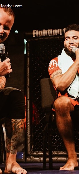 Two individuals are seated on a stage, each holding a microphone and smiling. They are in a sports-related setting with one wearing a casual black outfit and the other in sportswear, with a towel around his neck. The background includes elements of a fighting or martial arts cage.